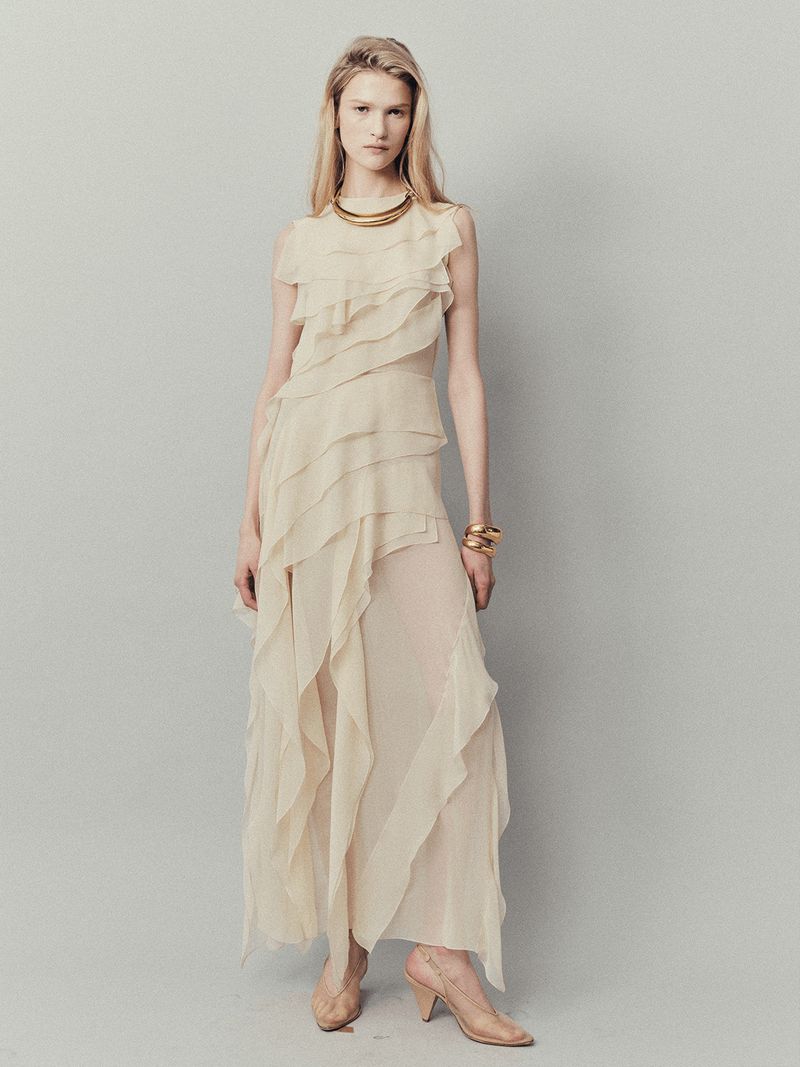 Full-length shot of a blond woman in a long beige chiffon dress with tiered ruffles, jewelry, and heels against a grey background.
