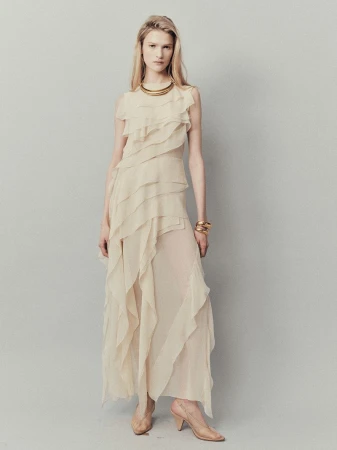 Full-length shot of a blond woman in a long beige chiffon dress with tiered ruffles, jewelry, and heels against a grey background.