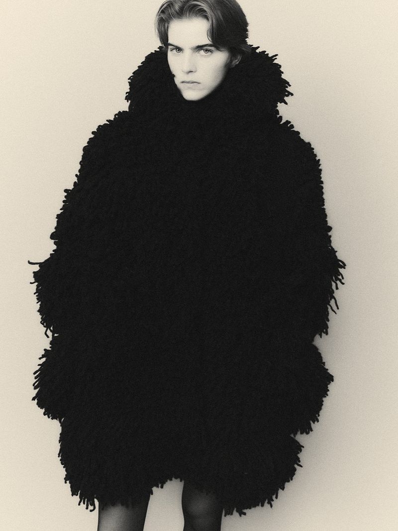 Black and white portrait of a fair-skinned person wearing a shaggy, textured black coat against a neutral backdrop in a studio setting.