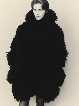Black and white portrait of a fair-skinned person wearing a shaggy, textured black coat against a neutral backdrop in a studio setting.