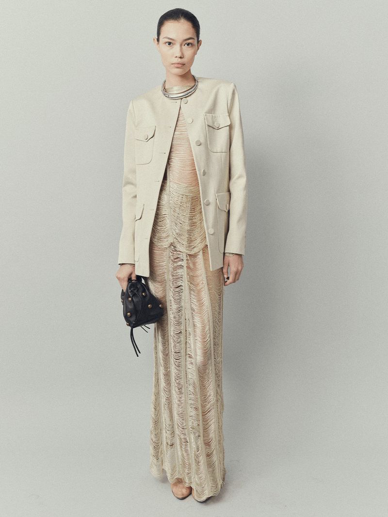 Full-length shot of a woman in a tan jacket and textured dress with a black purse in a neutral studio setting.