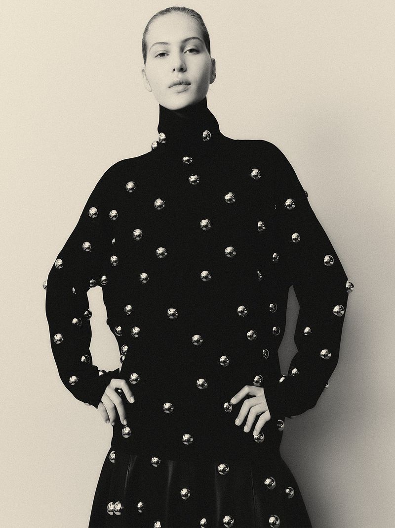 Elegant woman in a black, high-necked dress adorned with silver spheres, posing against a light neutral backdrop in a studio setting.