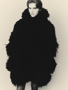 Model wearing an oversized voluminous black textured fringe coat with a high collar against a plain neutral studio background.