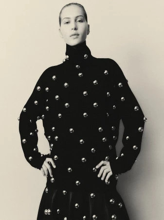 Model with slicked-back hair poses with hands on hips wearing a black turtleneck sweater covered in silver metallic sphere studs.
