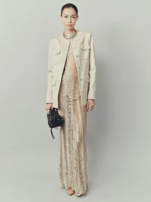 Model wearing a tailored beige jacket over a textured fringe gown with a silver choker and black bag against a grey backdrop.