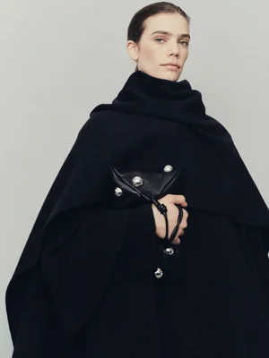 Model wearing a dark oversized wool coat holding a black leather handbag with silver hardware against a plain grey background