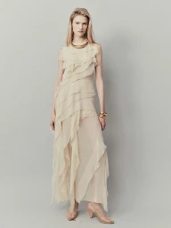 Model wearing a cream ruffled dress and gold statement jewelry against a plain grey background
