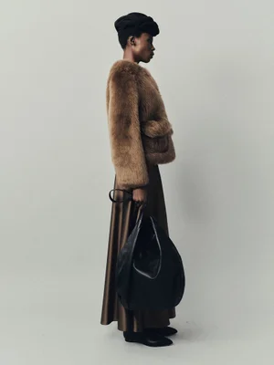 Model in side profile wearing a brown plush jacket, dark brown satin skirt, black headpiece, and holding a large black leather bag.