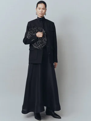 Model in black jacket and skirt holding a beaded handbag against a grey seamless backdrop for Altuzarra Fall 2025.