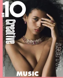 Close-up portrait of a woman wearing a jeweled necklace and rings against a grey background with magazine typography.