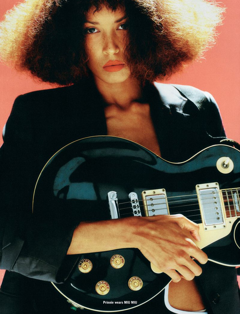 A woman with an afro is posing with a black electric guitar against a salmon-colored background in a fashion-style close-up portrait.