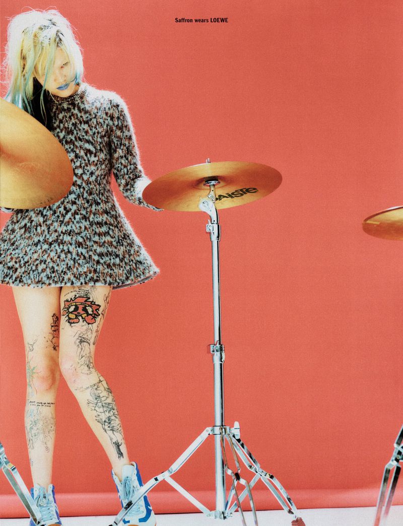 A tattooed woman in a patterned dress poses with drum cymbals against a red backdrop in a fashion editorial shot. The scene has a cool, edgy vibe.