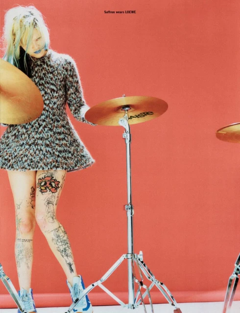 A tattooed woman in a patterned dress poses with drum cymbals against a red backdrop in a fashion editorial shot. The scene has a cool, edgy vibe.
