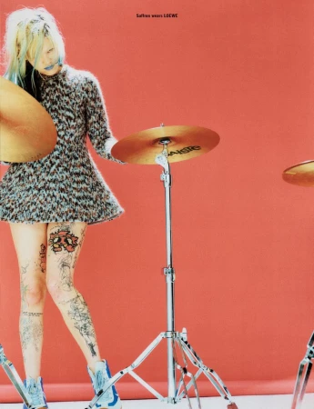 A tattooed woman in a patterned dress poses with drum cymbals against a red backdrop in a fashion editorial shot. The scene has a cool, edgy vibe.