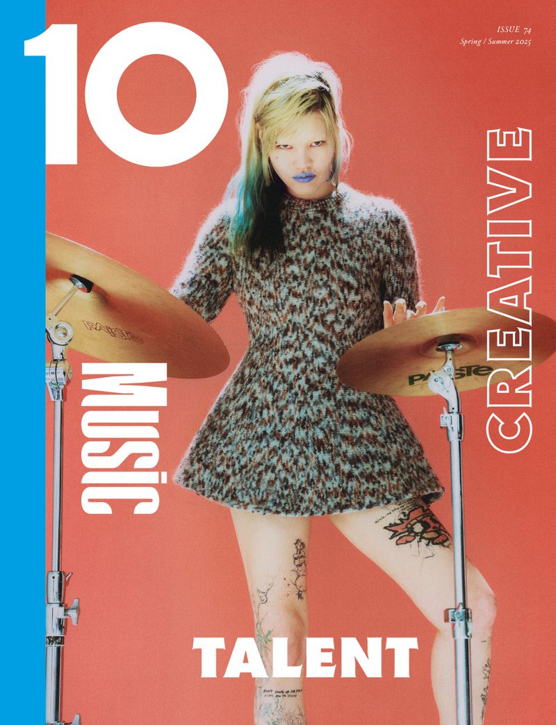 Magazine cover featuring a person in a patterned dress standing between drum cymbals against a coral-colored background with text overlay.
