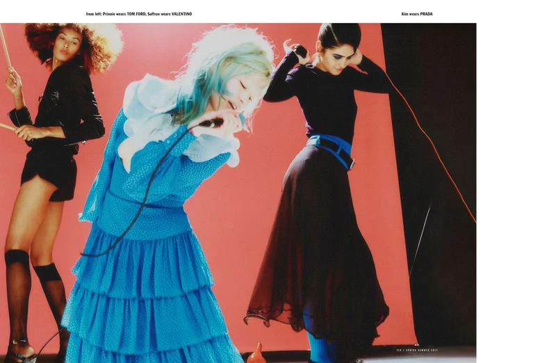 Three fashion models pose against a coral backdrop. One plays the drums in a blue dress, another in a black jacket and skirt.