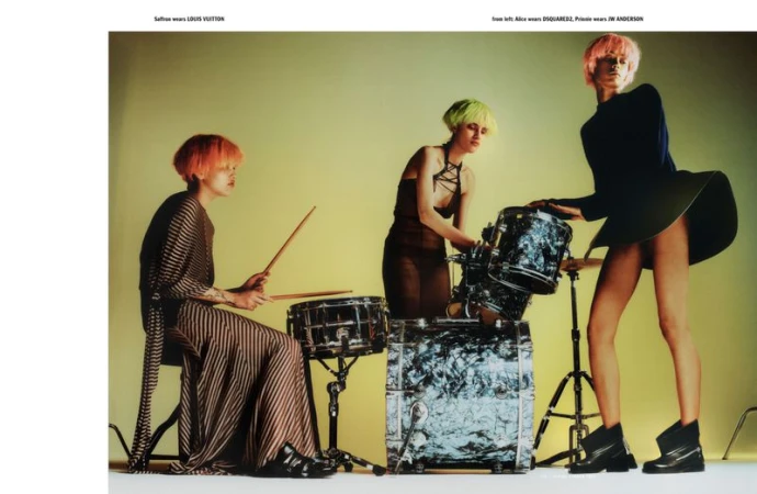 Three fashion models in an indoor photo studio setting, wearing designer dresses and playing a drum set with painted hair.