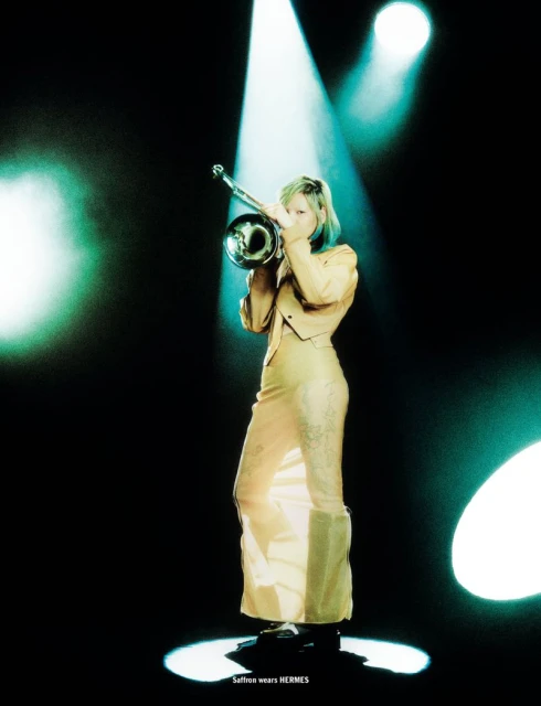 Woman in light yellow dress plays a trumpet under spotlights. Fashion or music editorial image with a dark background and stage lighting.