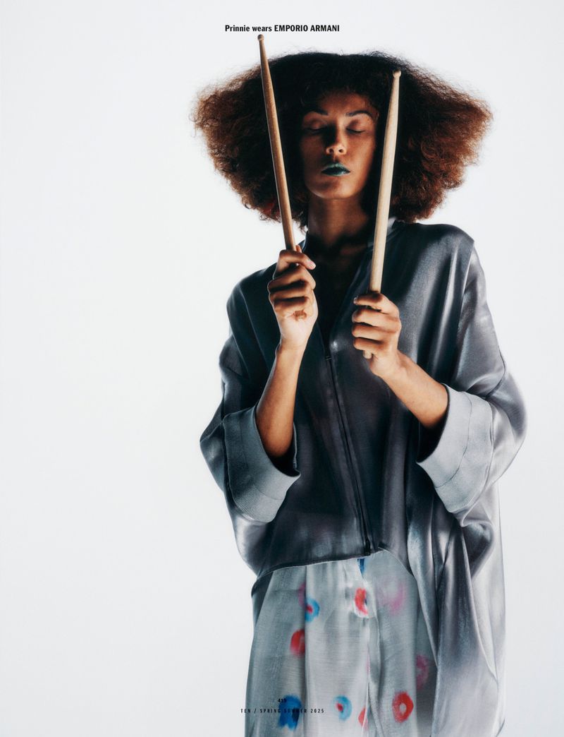 An adult female model with afro hairstyle holds drumsticks and wears a shiny grey jacket with printed skirt against a white backdrop.