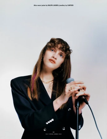Woman in dark blazer holding a microphone against a white background for 10 Magazine