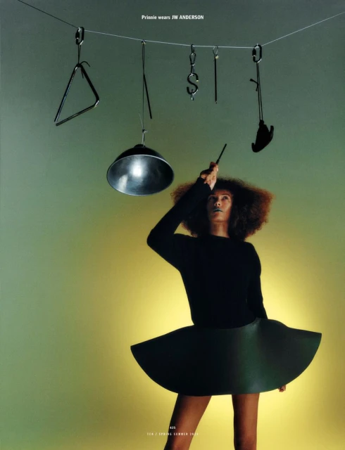 A woman with curly hair poses in a black outfit beneath hanging metal objects and lighting, creating a fashionable and artistic composition.