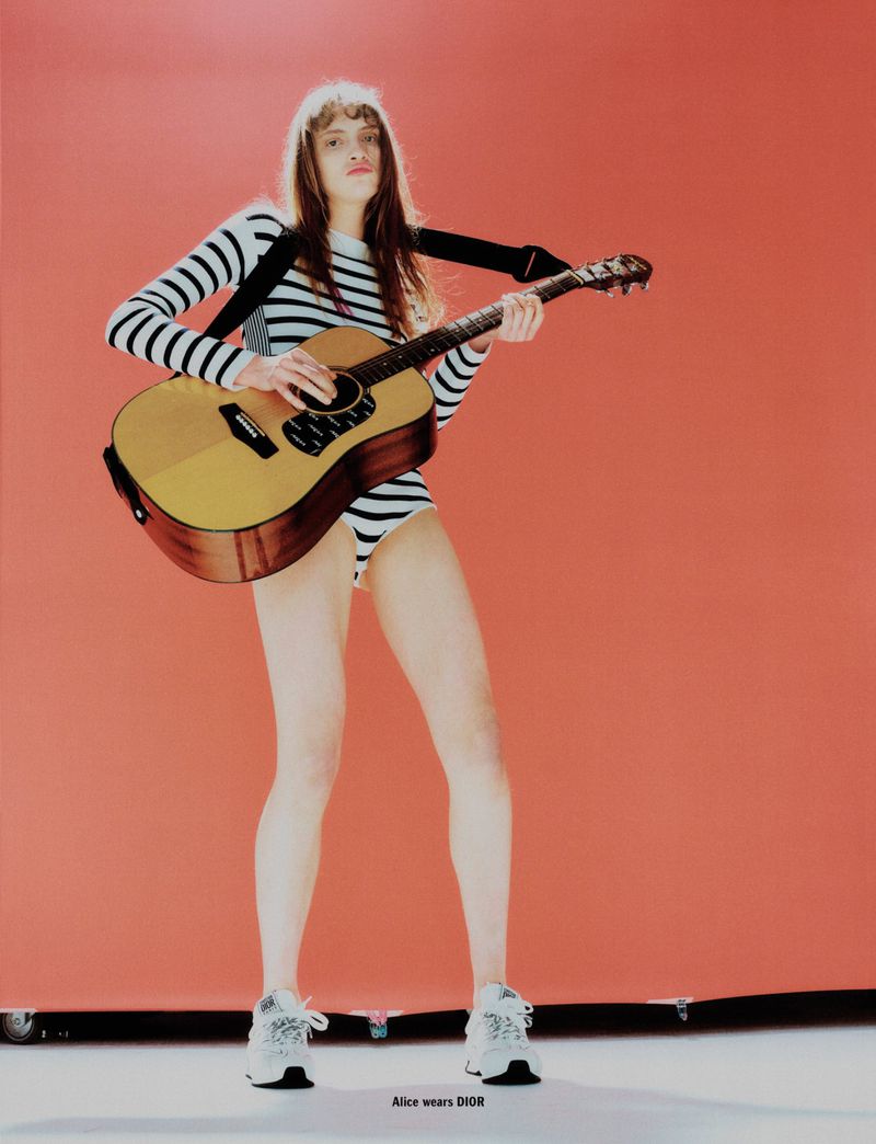 Young woman in striped bodysuit and sneakers poses with an acoustic guitar against a coral backdrop in a fashion editorial shot.