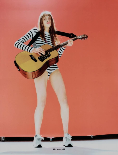 Young woman in striped bodysuit and sneakers poses with an acoustic guitar against a coral backdrop in a fashion editorial shot.