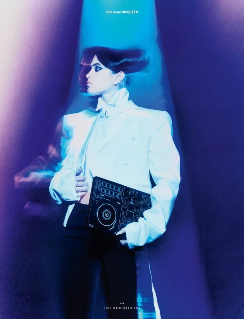 Stylish woman in white suit holds DJ controller in blue spotlight. Fashion editorial with modern aesthetic and high-fashion posing.