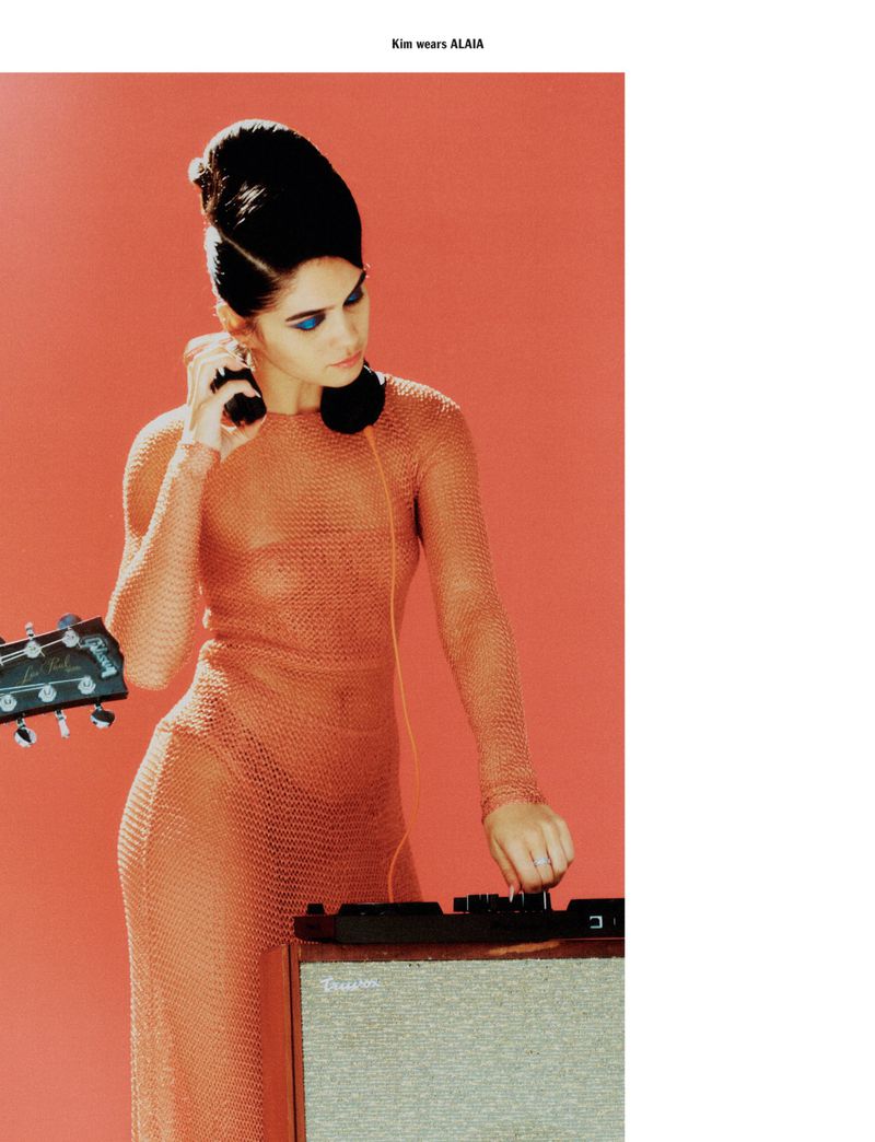 A woman in a sheer orange dress and vintage hair poses with a guitar and sound equipment against an orange backdrop in a studio setting.