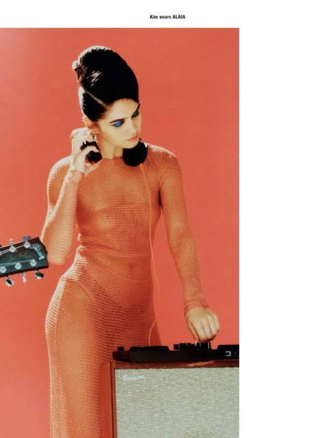 A woman in a sheer orange dress and vintage hair poses with a guitar and sound equipment against an orange backdrop in a studio setting.