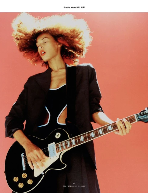Editorial shot of a Black female model playing an electric guitar against a coral background, wearing a black suit and Miu Miu outfit.
