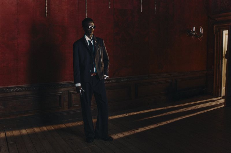 An African-American adult male model in a dark suit and sunglasses stands in a dimly lit room with red walls and wooden floors.