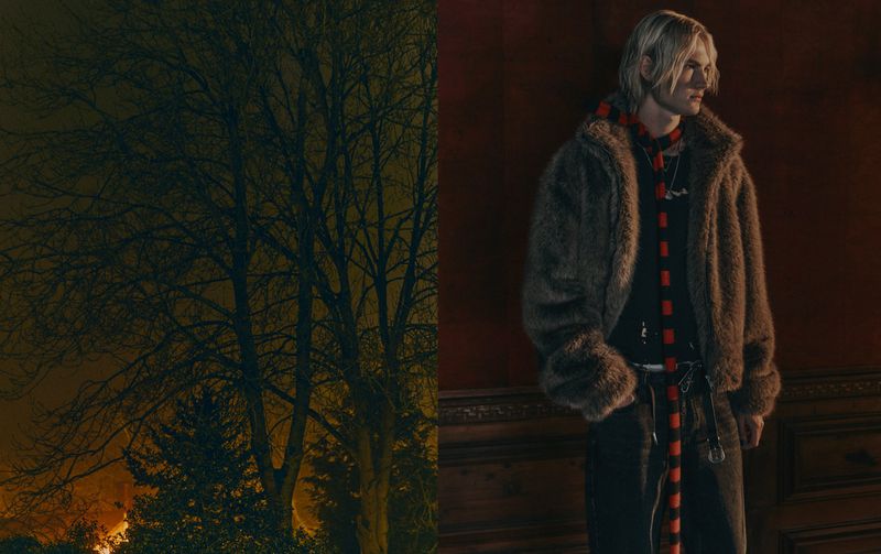 A man with blond hair, wearing a fur coat and scarf, stands against a red wall with dark wood trim, with a tree in the background.