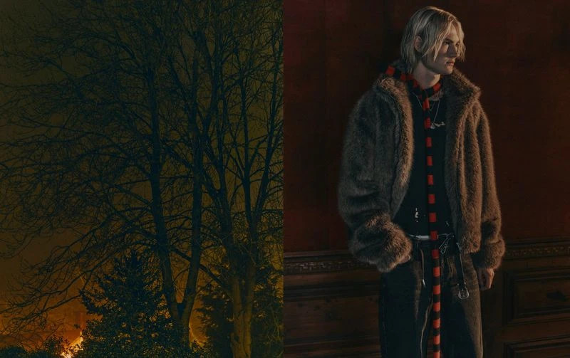 A man with blond hair, wearing a fur coat and scarf, stands against a red wall with dark wood trim, with a tree in the background.