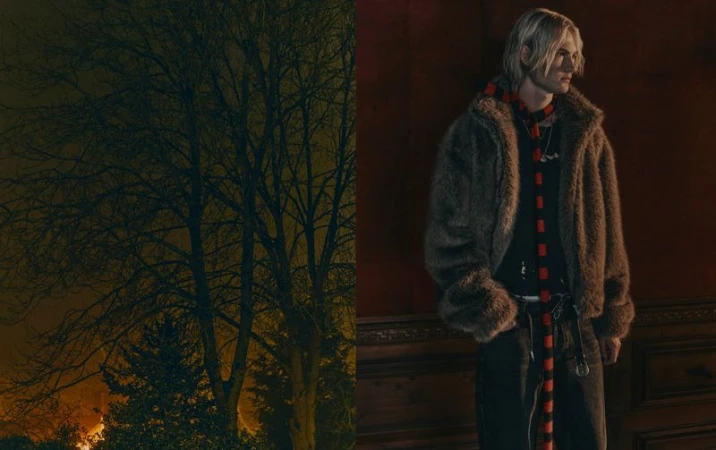 A man with blond hair, wearing a fur coat and scarf, stands against a red wall with dark wood trim, with a tree in the background.