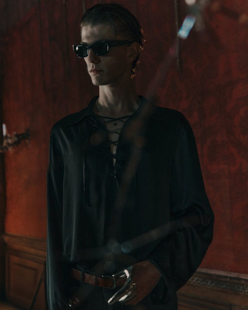 Portrait of a slender adult male wearing sunglasses and a lace-up black silk shirt, standing indoors against a textured red wall.