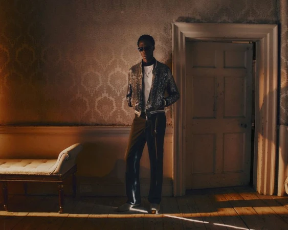 Cherif Douamba posing in a sequined bomber jacket, dark trousers, and sunglasses in a dimly lit room with vintage wallpaper.