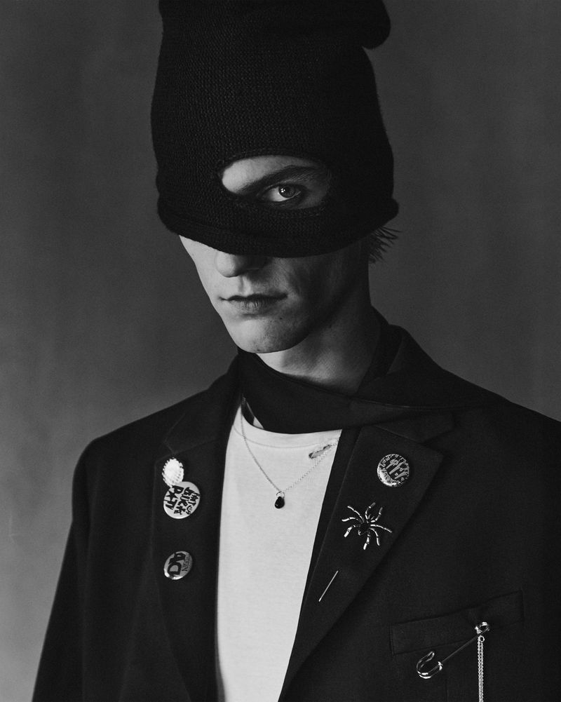 Monochrome portrait of a person in a black ski mask and dark blazer adorned with pins and a necklace against a grey backdrop.
