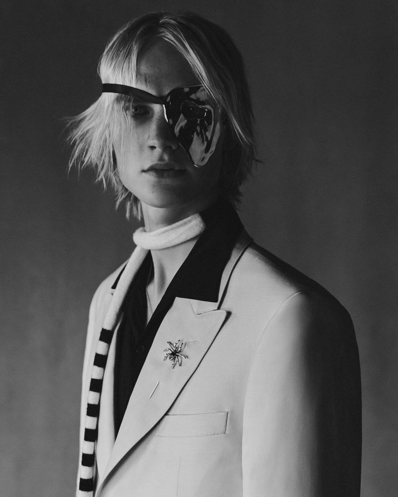 Black and white studio portrait of a young adult with blond hair, an eye patch, and a light colored suit with brooch detail.