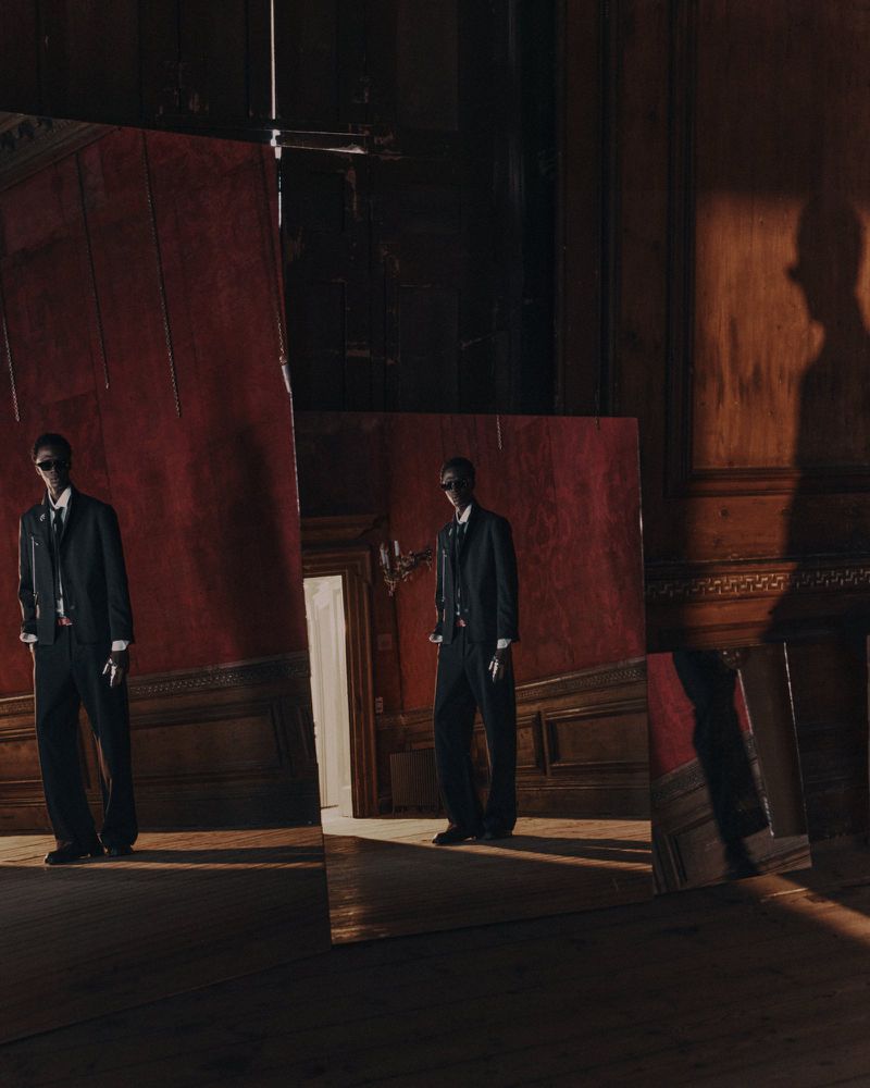 Adult male wearing a black suit and sunglasses stands indoors reflected in mirrors on a hardwood floor in a fashion-style editorial shot.