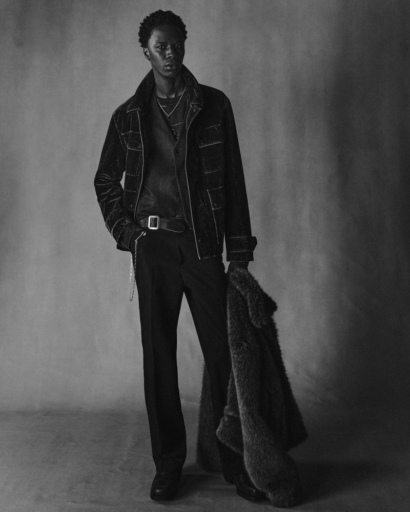 Cherif Douamba posing in a dark topstitched jacket and black trousers holding a heavy coat against a mottled backdrop.