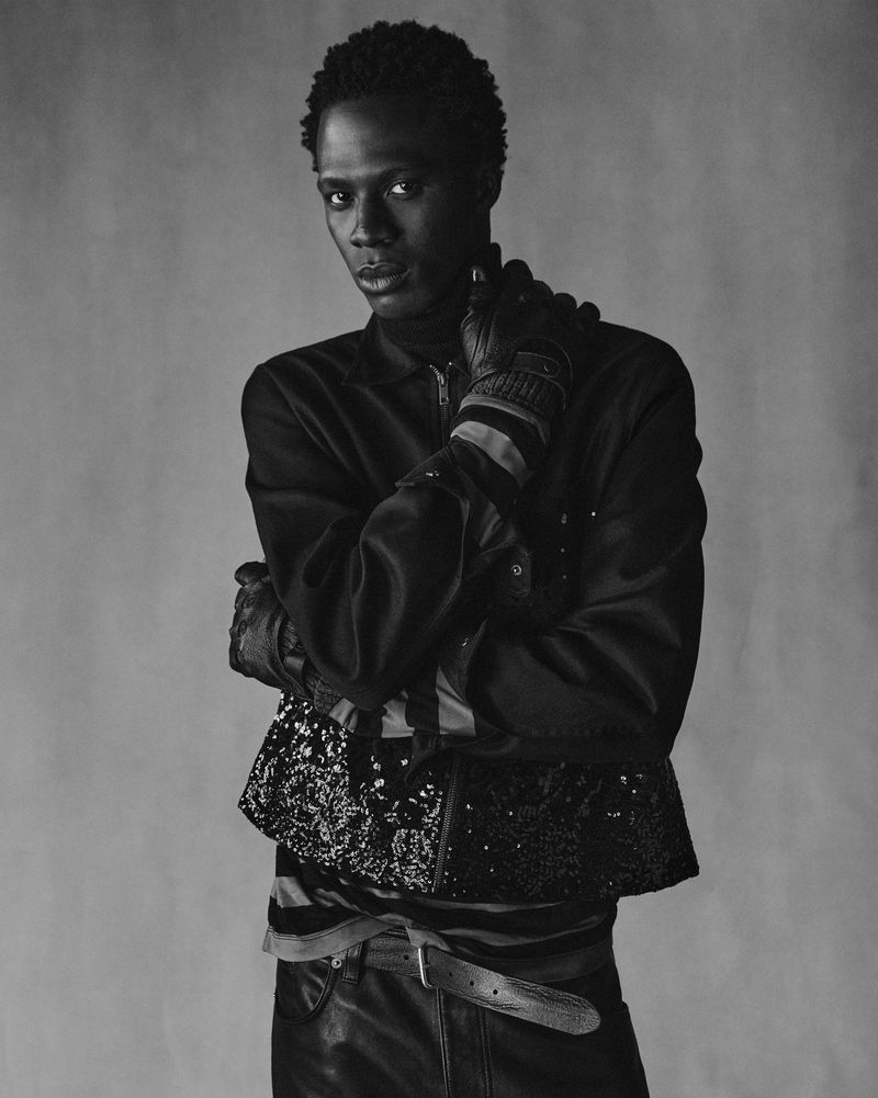 Black and white portrait of Cherif Douamba wearing a layered jacket with sequin trim and leather gloves against a textured studio backdrop.
