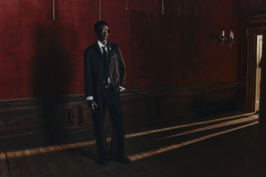 Model wearing dark suit jacket and trousers in a wood-paneled room with deep red walls and light streaming across the floor