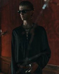 A man wearing dark sunglasses and a black lace-up shirt posing against a dark red wall in a moody interior.