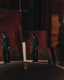 A man in a dark suit and sunglasses standing in a dim room with red wallpaper, reflected across multiple leaning floor mirrors.
