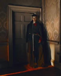 Male model wearing a layered dark coat, striped top, and wide-leg trousers standing near an interior door with vintage wallpaper.