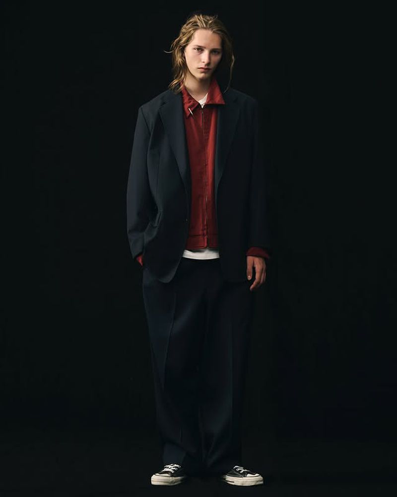 Full-length studio shot of a young adult male model in a dark blazer, red shirt, and dark pants against a black backdrop.