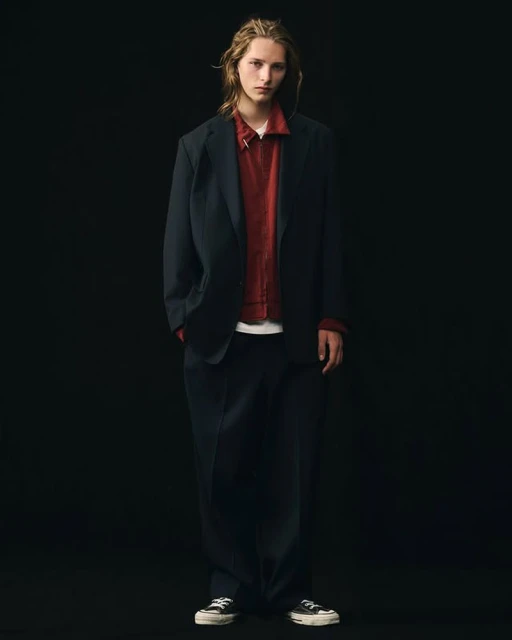 Full-length studio shot of a young adult male model in a dark blazer, red shirt, and dark pants against a black backdrop.