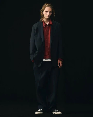 Full-length studio shot of a young adult male model in a dark blazer, red shirt, and dark pants against a black backdrop.