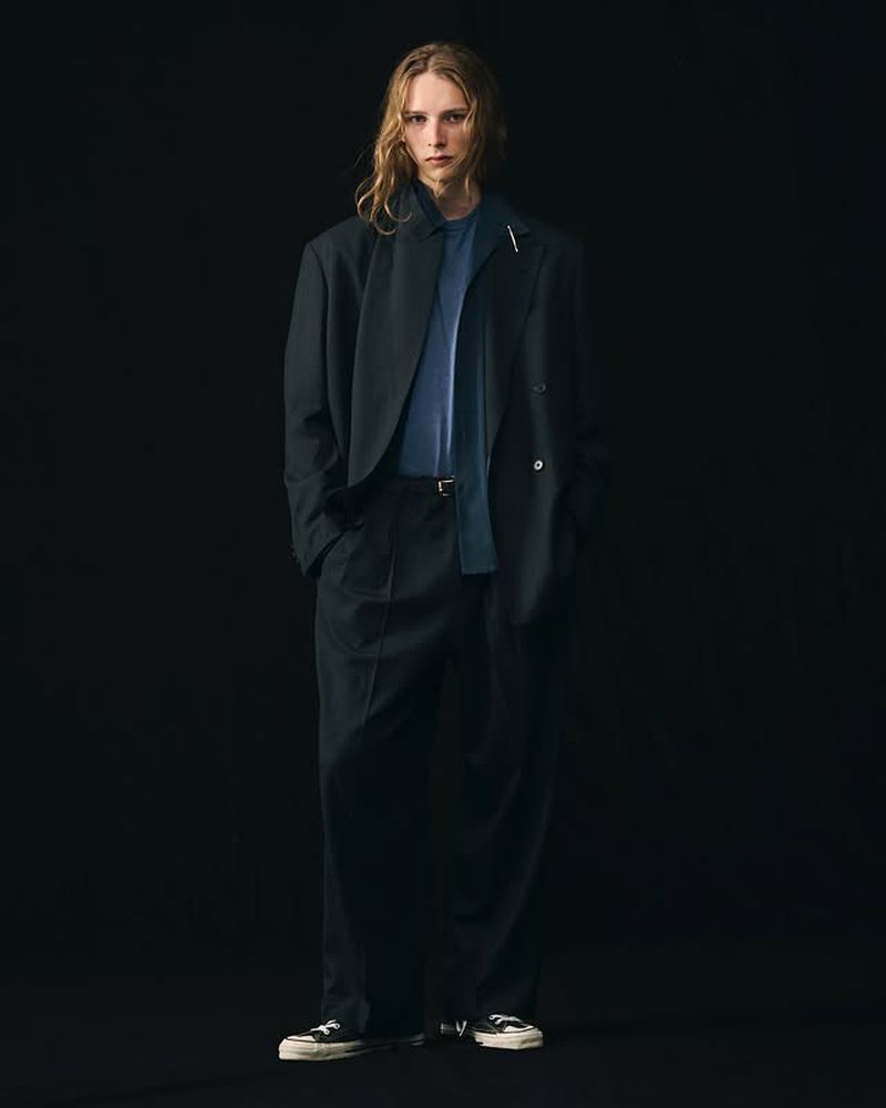 Adult male model in a relaxed pose wearing a dark blazer, blue tee shirt, and wide leg pants, photographed indoors with dark background.